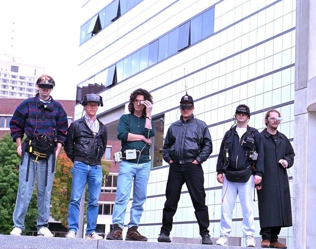 Six members of the MIT Media Lab wearable computing group standing outside the Media Lab in the 1990s, wearing head-mounted displays and belt-bag computers.